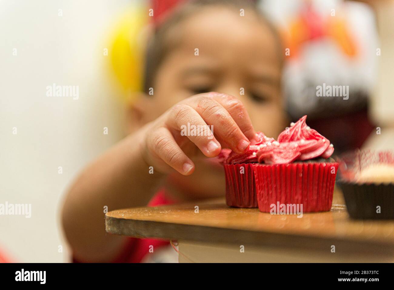 Blurred image of a boy picking up a creamy, red cupcake. Child enjoying sweet on children's birthday. Child smearing himself with a chocolate cupcake Stock Photo