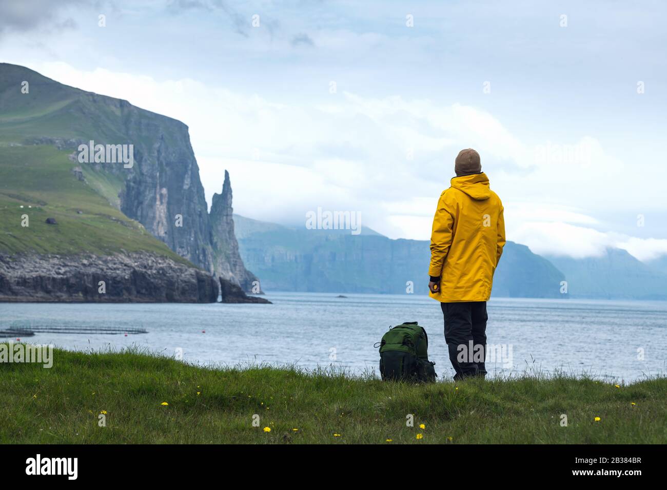 Tourist in yellow jacket looks at Witches Finger cliffs from Trollkonufingur viewpoint. Vagar island, Faroe Islands, Denmark. Stock Photo