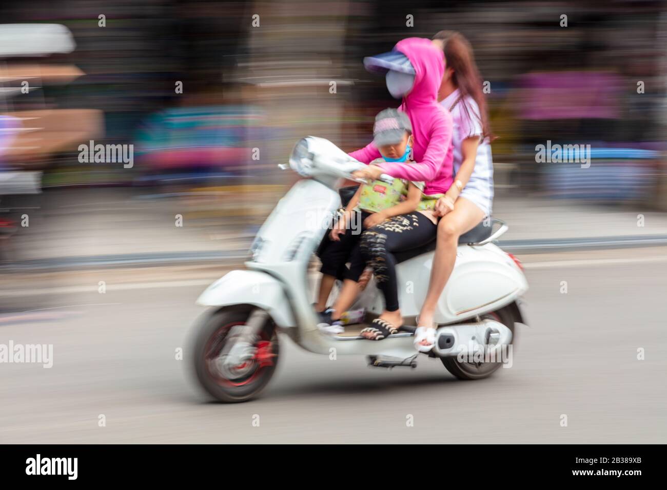 Motion blurred photograph of local people, women and a child, wearing facemasks, riding moped scooter on the streets of Hanoi, Vietnam, April 3 2018 Stock Photo