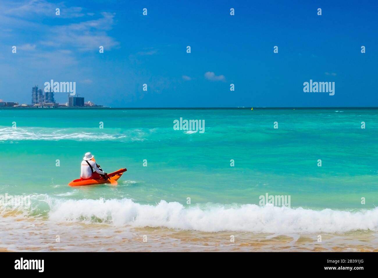 Lifeguard on duty seen from behind in orange boat and paddle in hand going into sea. Stock Photo