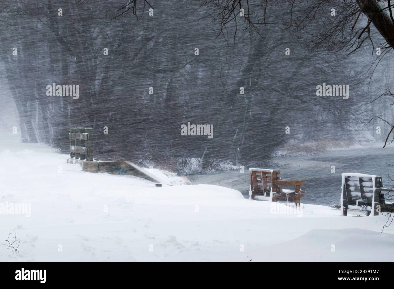 Snow blowing sideways over a pond and a bridge with a park bench on the side. Stock Photo
