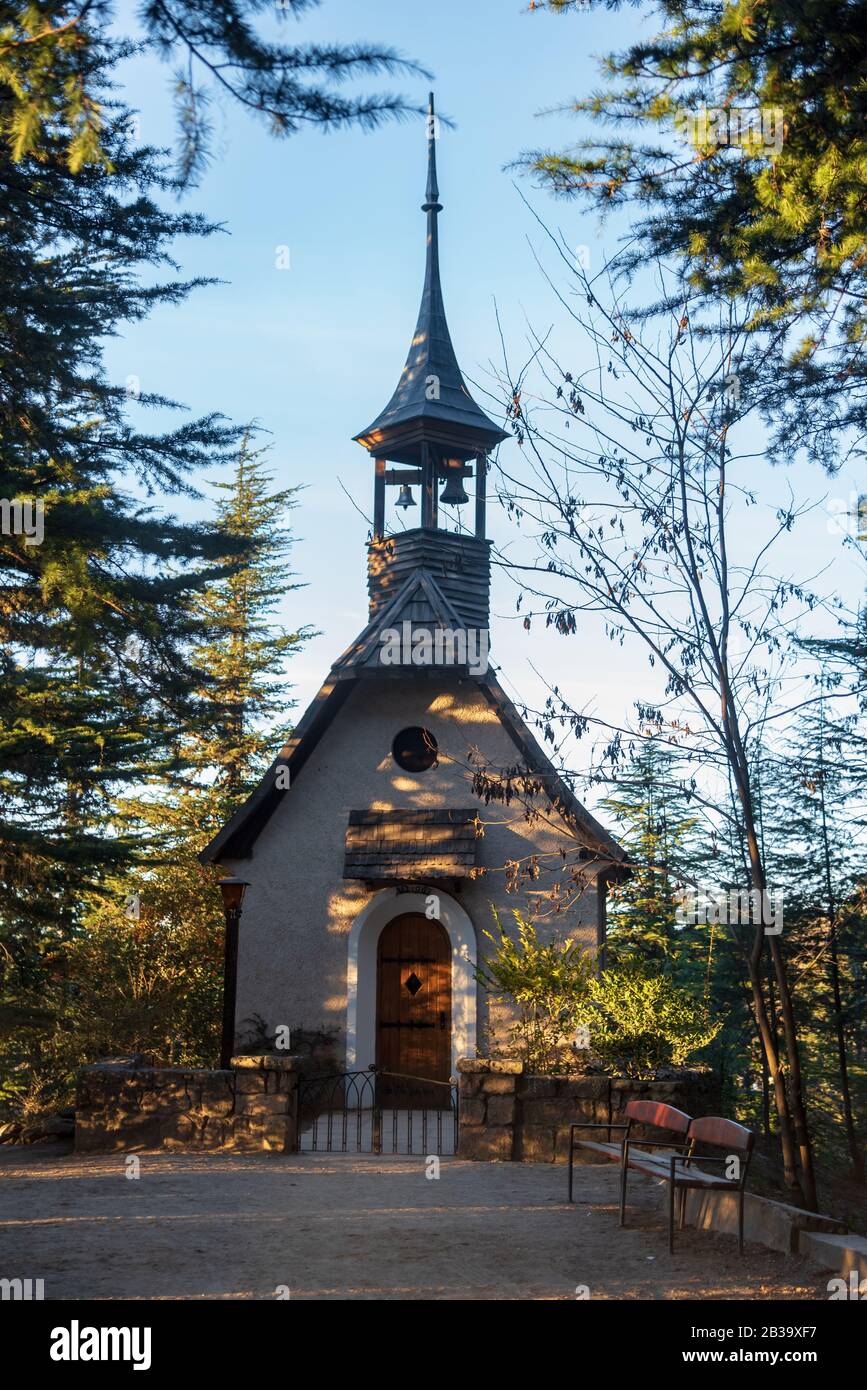 Germanic small wooden church with two bells at the tower in La Cumbrecita, with a dirt road and a bench at the front of it, surrounded by evergreen tr Stock Photo
