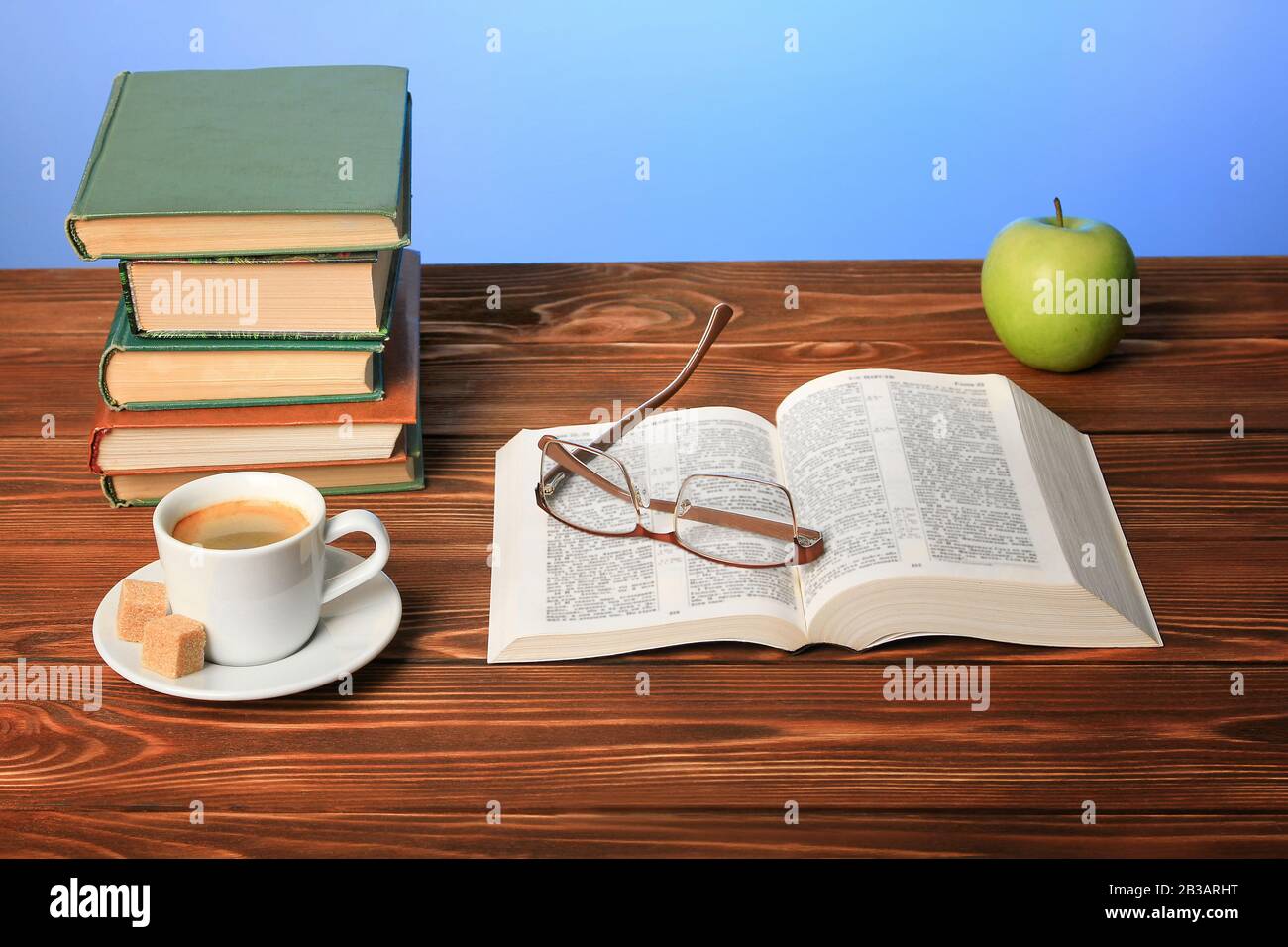 An open book on the table, a green apple, glasses, a cup of cappuccino coffee with sugar cubes, blue background. Stock Photo