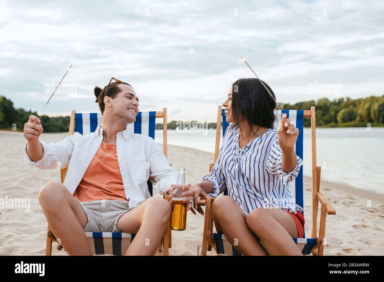 cheerful interracial couple sitting in chaise lounges and holding sparklers Stock Photo