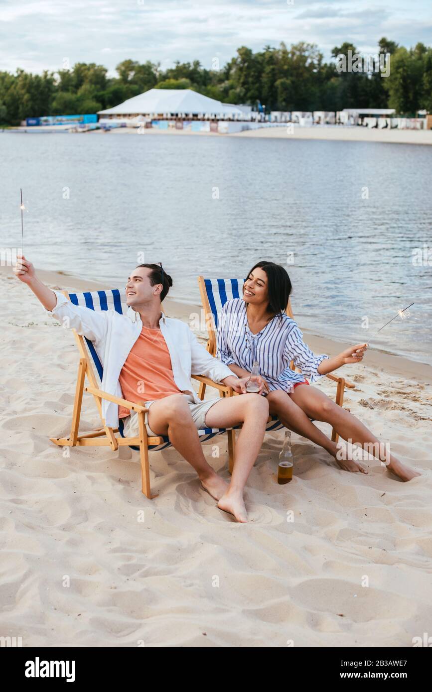 happy interracial couple sitting in chaise lounges and holding sparklers while resting on beach Stock Photo