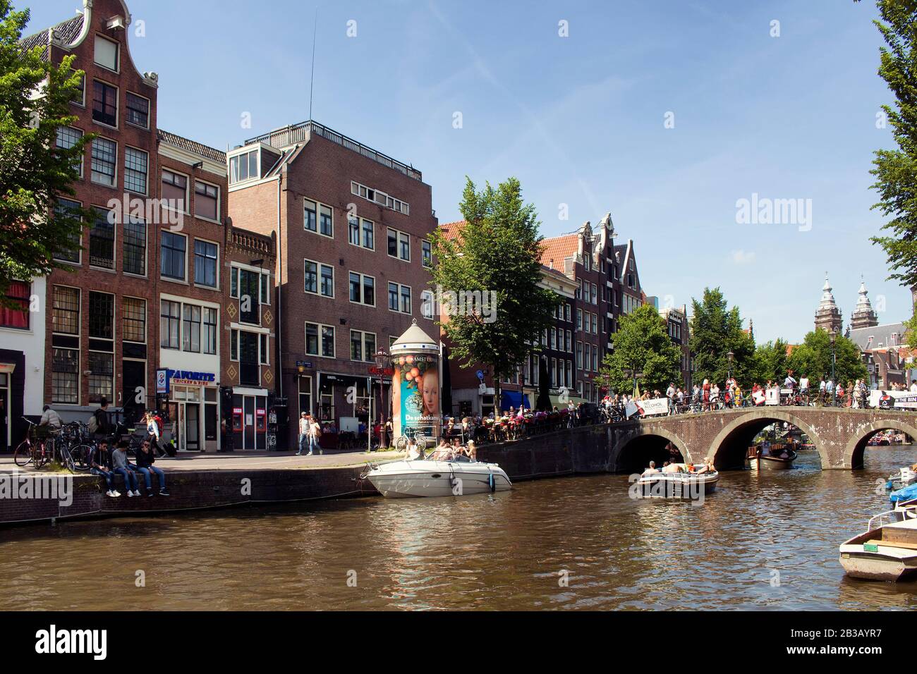 View of people riding a boat on canal doing a cruise tour in Amsterdam. Many people hang out on street and crossing Armbrug bridge. It is a sunny summ Stock Photo