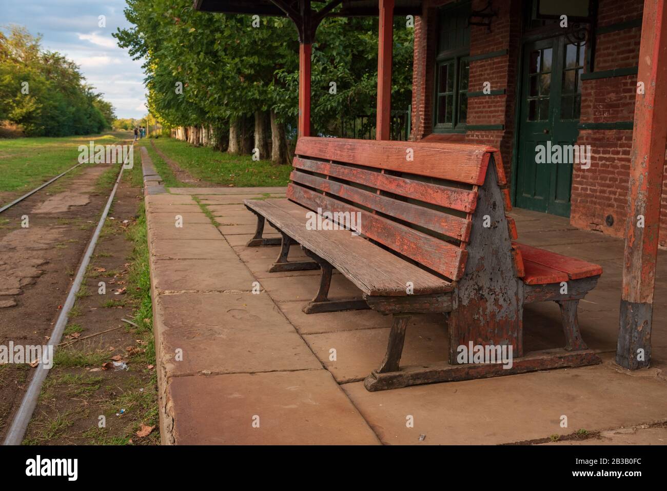 A wooden bench in an old abandoned train station with a view of the railroad with grass aroun it Stock Photo