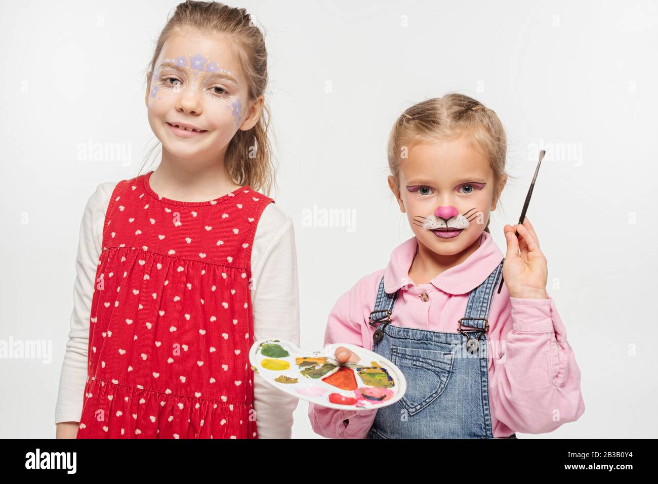 adorable kid with cat muzzle painting of face holding palette and paintbrush near friend with painted floral mask isolated on white Stock Photo