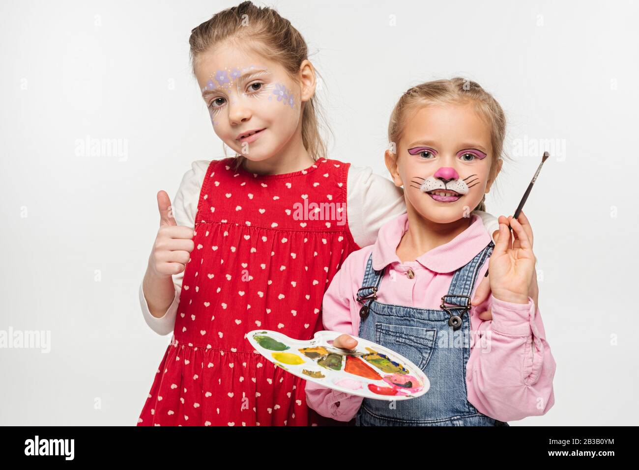 adorable kid with cat muzzle painting of face holding palette and paintbrush, while friend with floral mask showing thumb up isolated on white Stock Photo