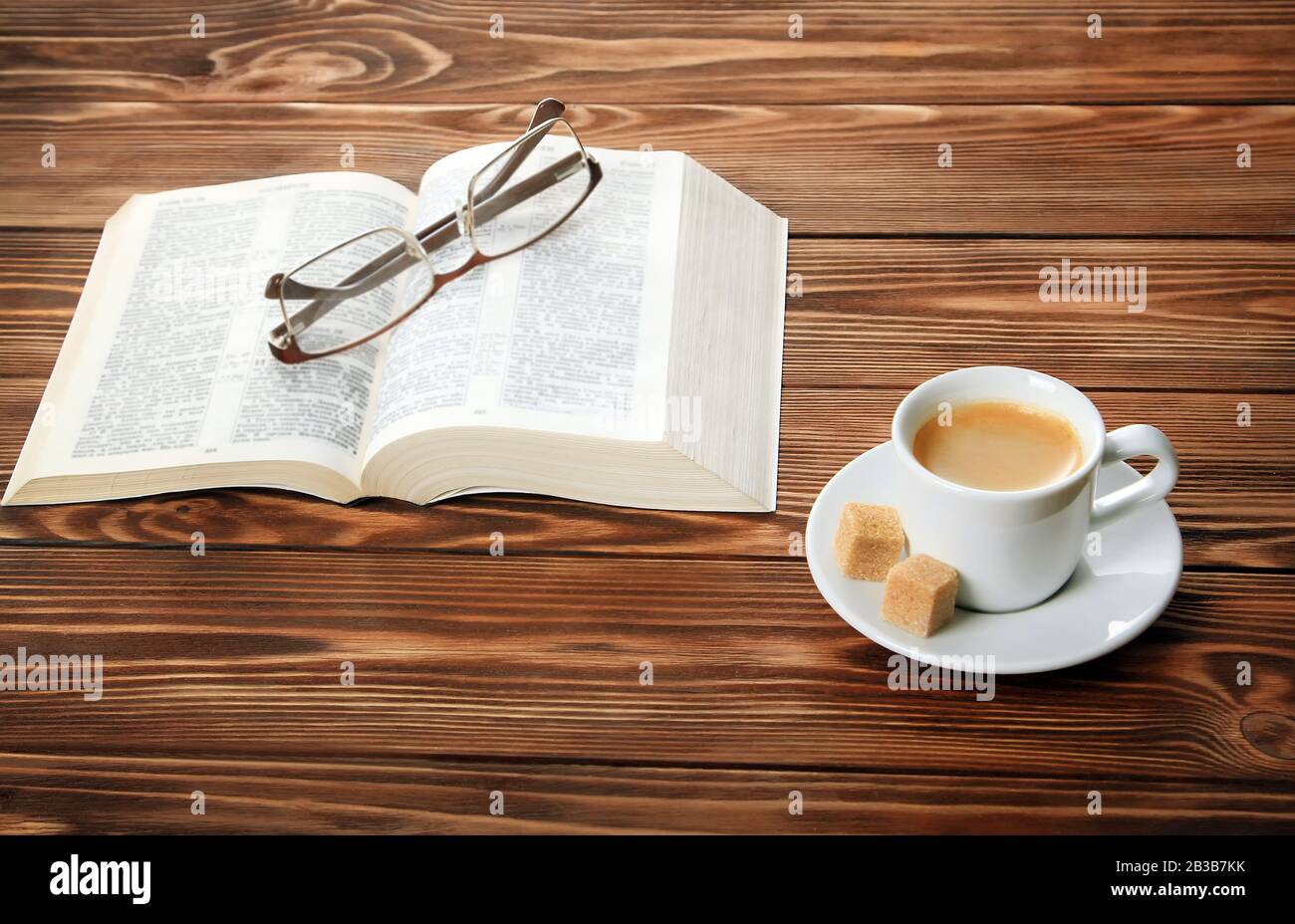 An open book on the table, glasses, a cup of cappuccino coffee with sugar cubes, blue background. Stock Photo