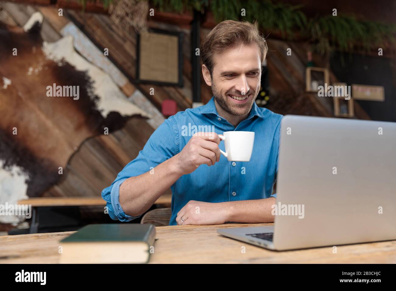 Joyful casual man reading, laughing, and drinking his coffee while wearing a blue shirt, sitting at a desk on coffeeshop background Stock Photo
