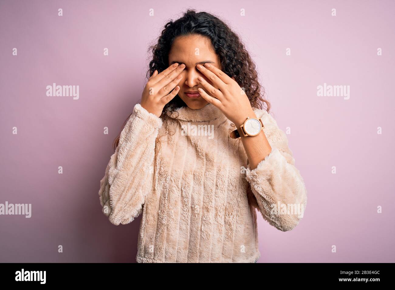 Young beautiful woman with curly hair wearing casual sweater standing over pink background rubbing eyes for fatigue and headache, sleepy and tired exp Stock Photo