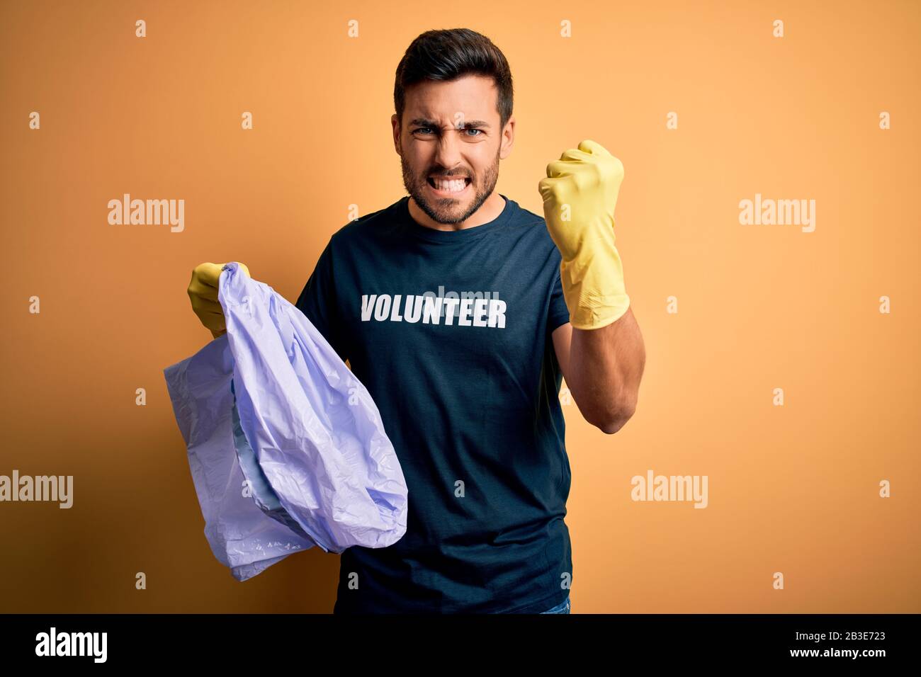 Young handsome volunteer man with beard cleaning junk using bag over yellow background annoyed and frustrated shouting with anger, crazy and yelling w Stock Photo