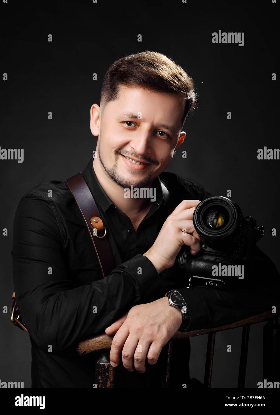 Happy man photographer with photo camera on black background. Portrait of smiling male model in black fashion clothes taking photos in studio Stock Photo