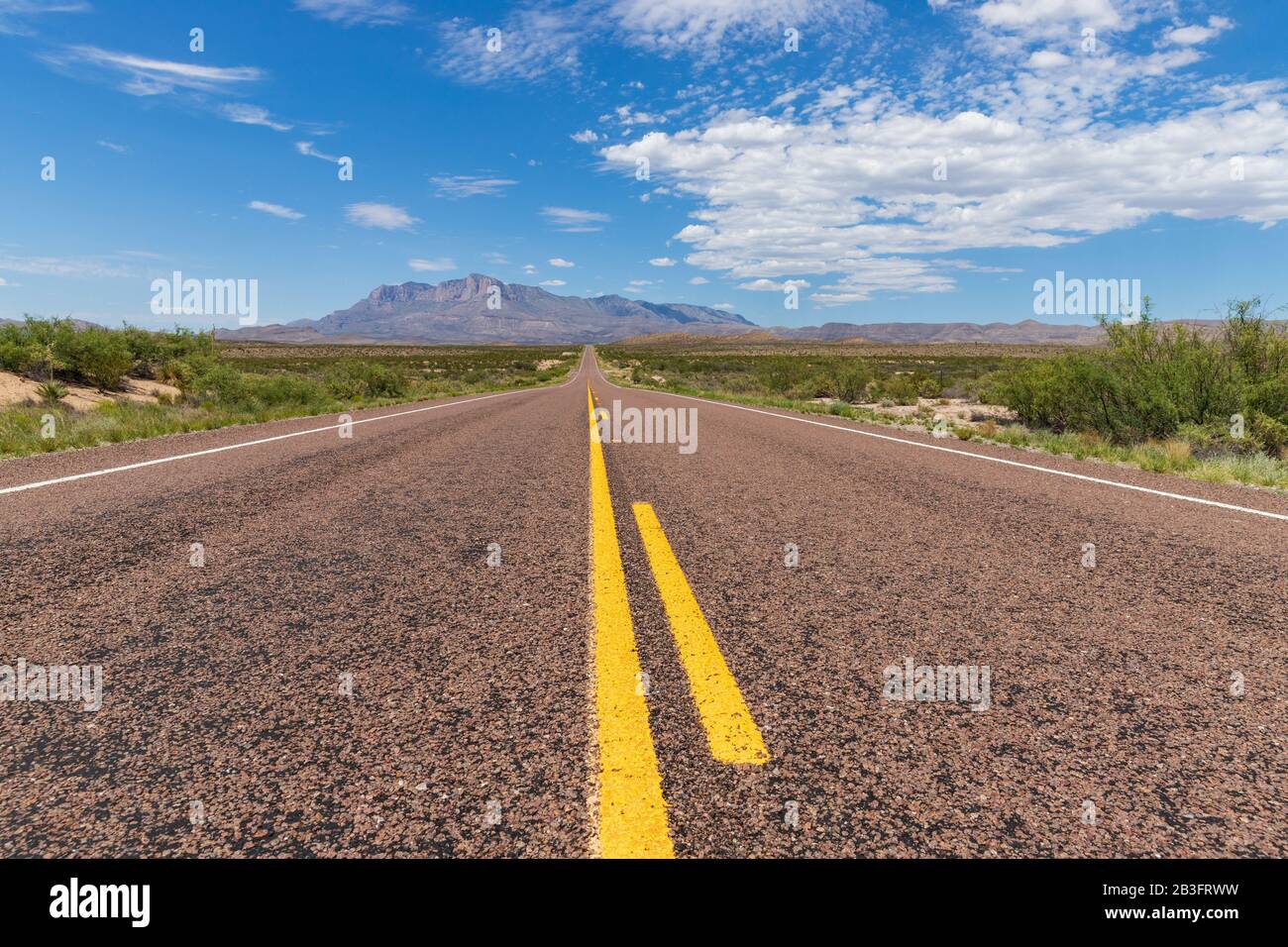 Long straight road in the desert, leading to a beautiful mountain range under a blue sky with clouds Stock Photo