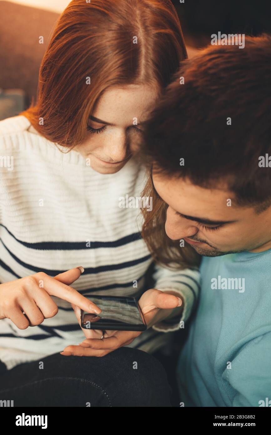 Red haired caucasian girl with freckles embraced by her lover is looking for something in her mobile phone Stock Photo