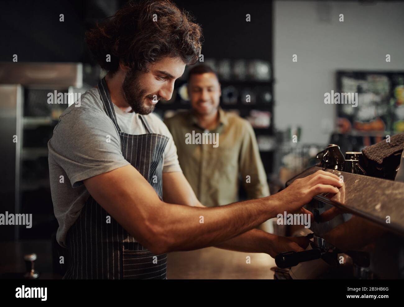 Handsome successful male worker making fresh coffee using machine while customer watching through counter Stock Photo