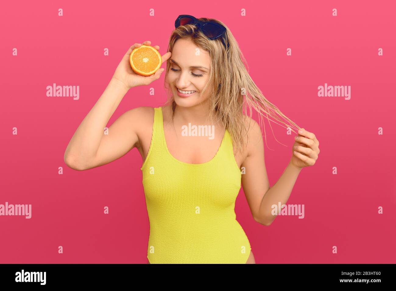 Happy healthy young woman holding a halved fresh juicy orange to her eye with a beaming friendly smile over a pink studio background Stock Photo