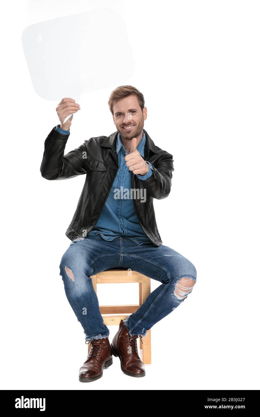 beautiful casual man with black leather jacket is sitting on a wooden chair holding a speech bubble overhead with a ok sign gesture on white studio ba Stock Photo