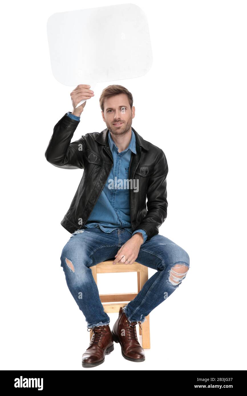 fine casual man with black leather jacket is sitting on a wooden chair holding a speech bubble overhead happy on white studio background Stock Photo