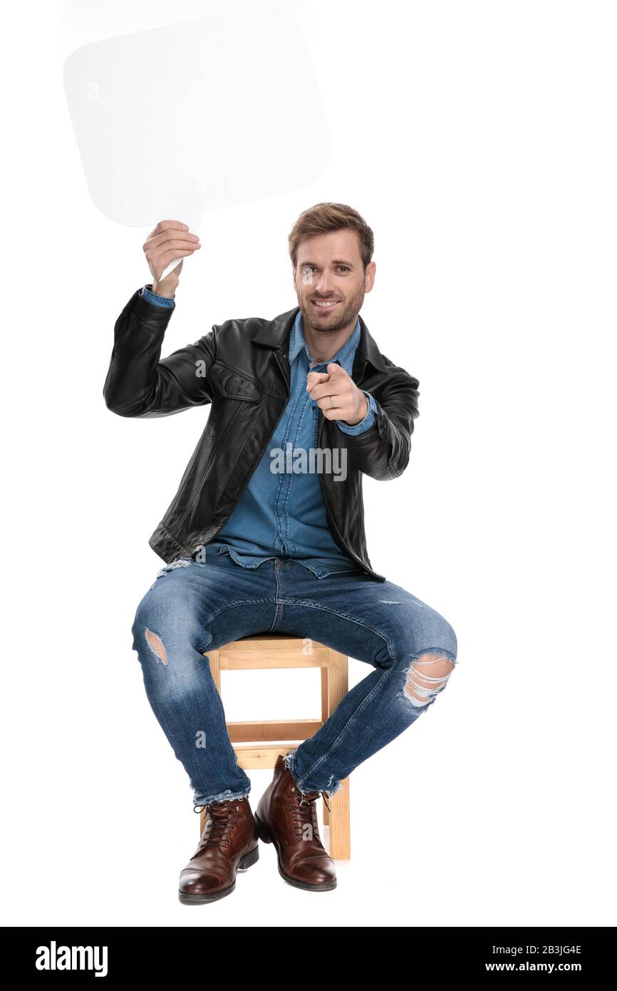 attractive casual man with black leather jacket is sitting on a wooden chair holding speech bubble overhead and pointing forward happy Stock Photo