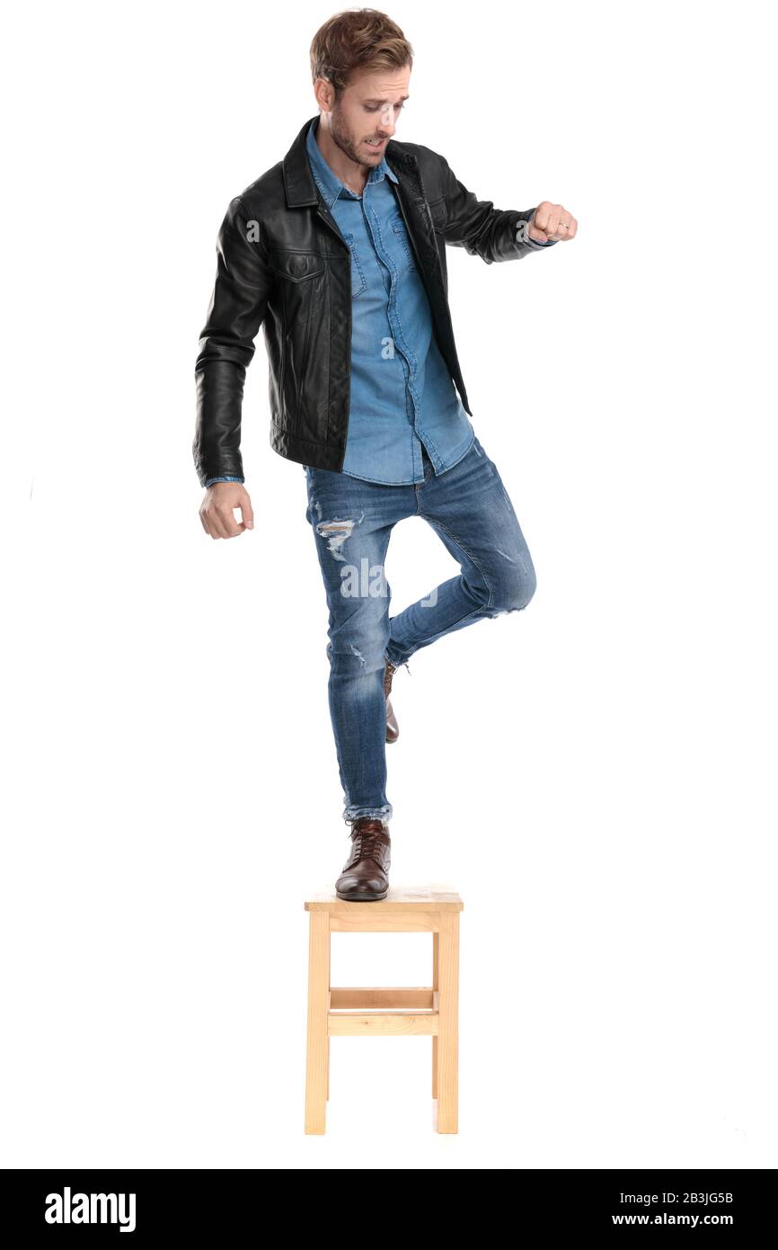 handsome casual man with black leather jacket mantaining his balance avoiding a fall off the chair in one leg scared on white studio background Stock Photo