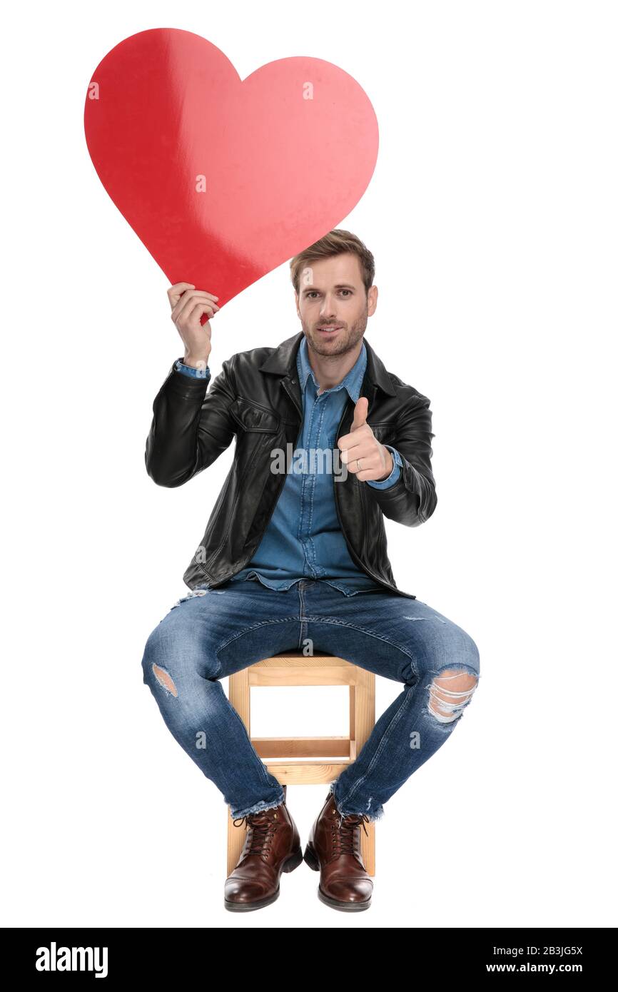 charming casual man with black leather jacket is sitting on a wooden chair holding a big red heart overhead with a ok sign  on white studio background Stock Photo
