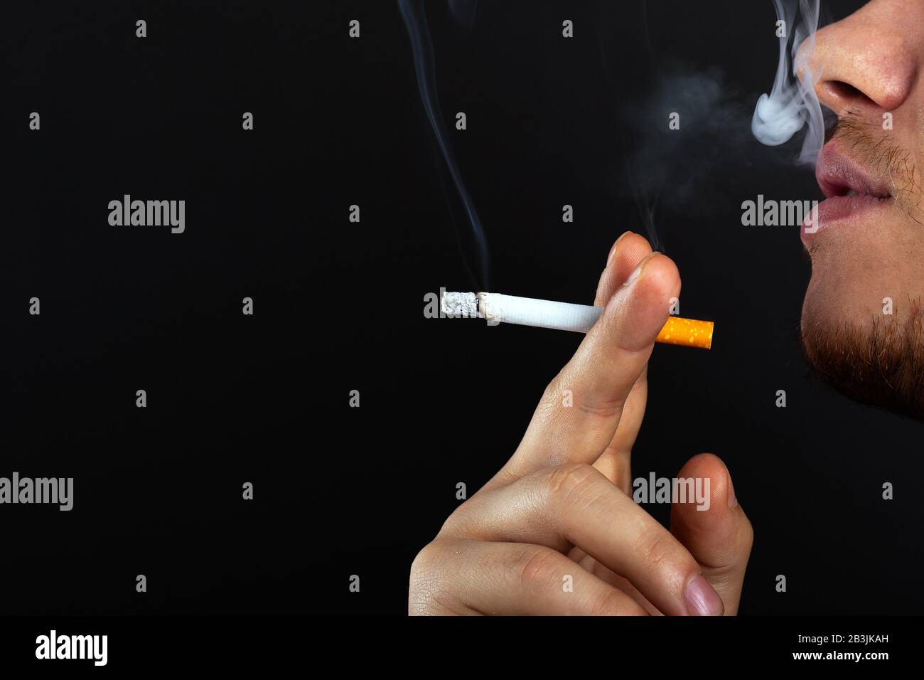 A young man holds a cigarette in his hands and exhales smoke on a dark background. harm from smoking nicotine and tobacco use close-up. Stock Photo