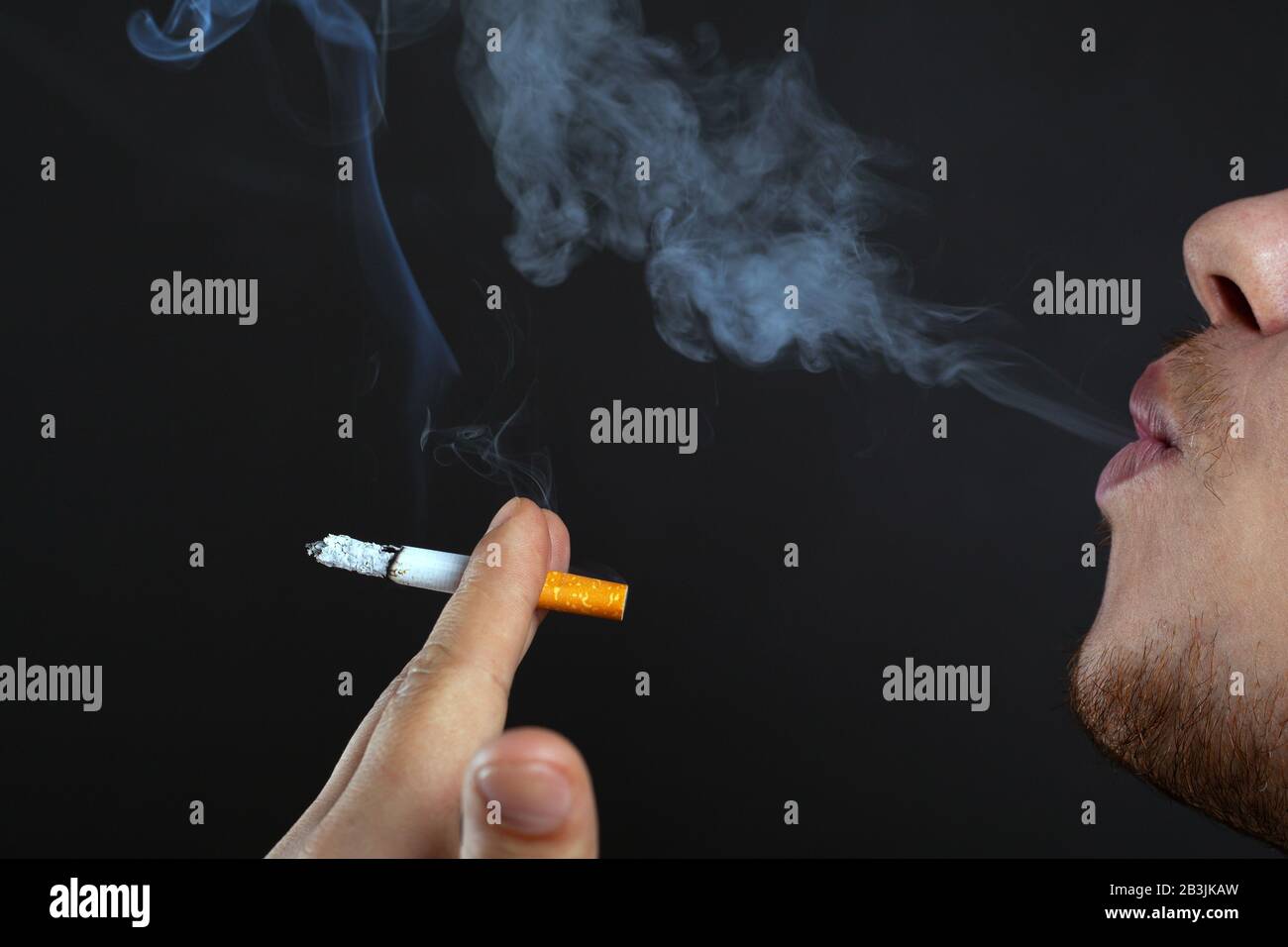 young man holding a smoking cigarette in his hands and exhales smoke on a dark background. harm from nicotine and tobacco close-up. Stock Photo