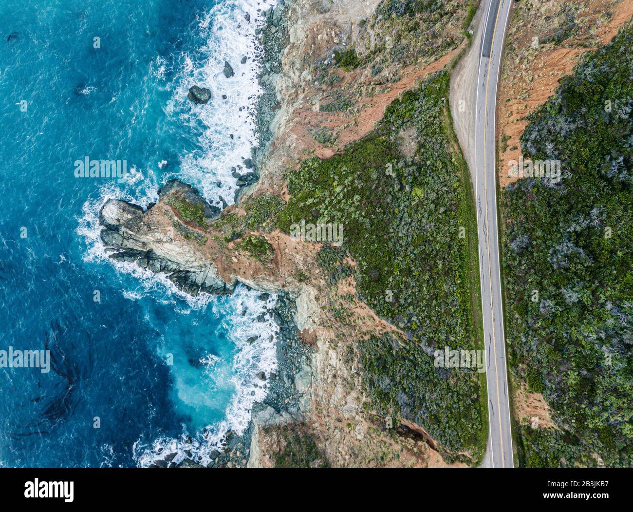 Aerial view of the waves breaking on the rock beside a road. Light and dark blue water foam while the waves break on the shore. Deep sea. Big Sur Cali Stock Photo
