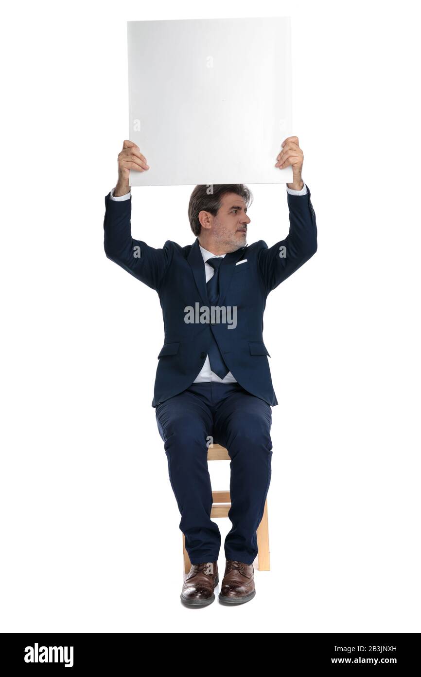 good looking formal business man with navy suit is sitting and holding a billboard overhead while looking to a side happy on white studio background Stock Photo