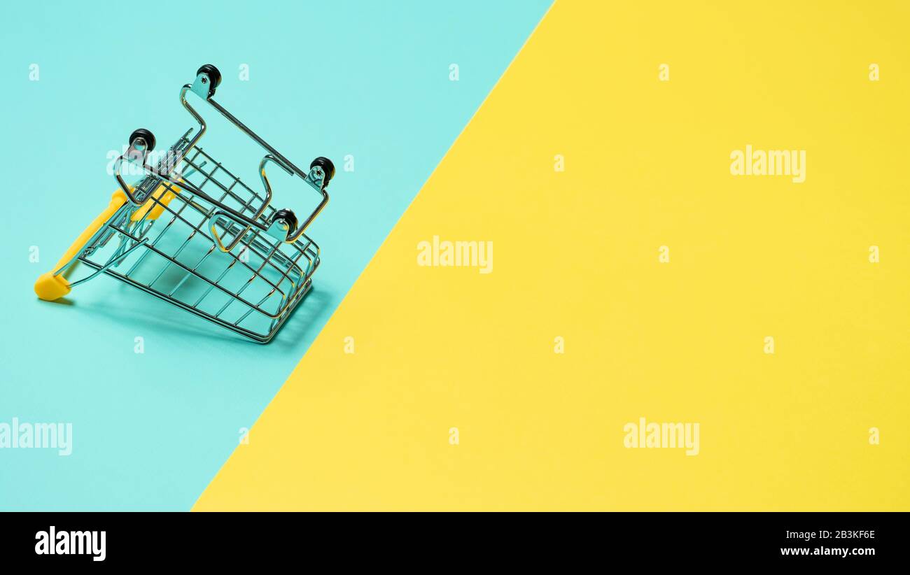 Empty inverted shopping cart on blue and yellow background. Inverted toy trolley on bright colorful background, copy space for text or design. Stock Photo