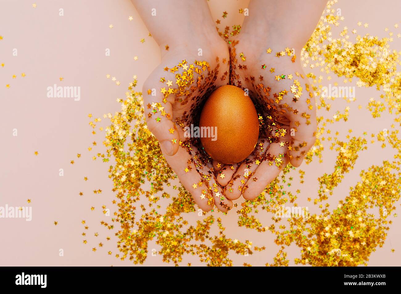 light skin hand in gold spangles in the form of stars firmly hold a Golden Easter egg on a pink background with sequins. Happy Easter flat lay mockup Stock Photo