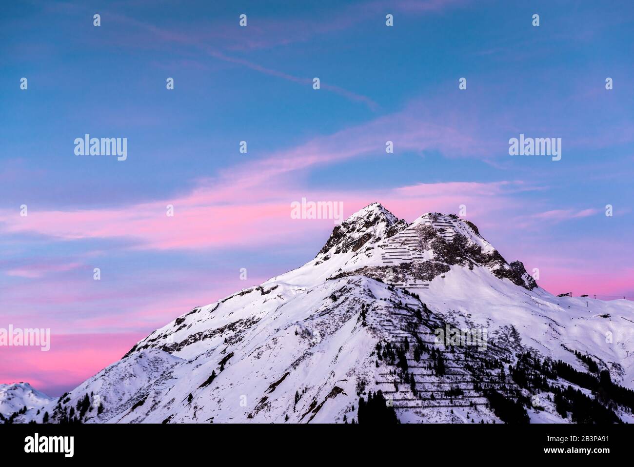 Pink clouds lie in a blue sky above a snowy alpine mountain peak. Stock Photo
