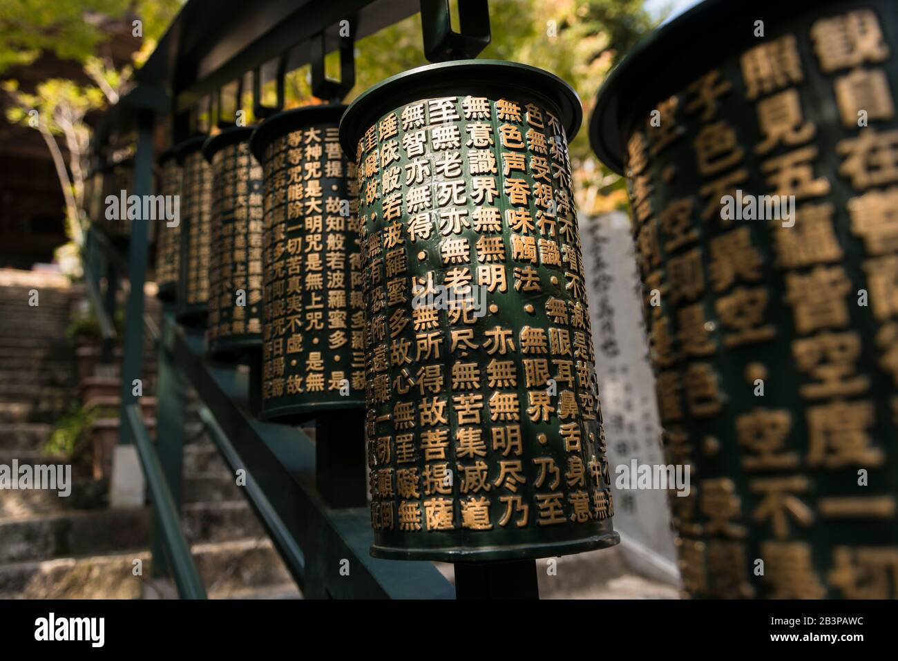 Spinning cylindrical metal prayer wheels with golden lettering beside steps to a temple within Daisho-in Temple complex, Mount Mison, Miyajima Island Stock Photo