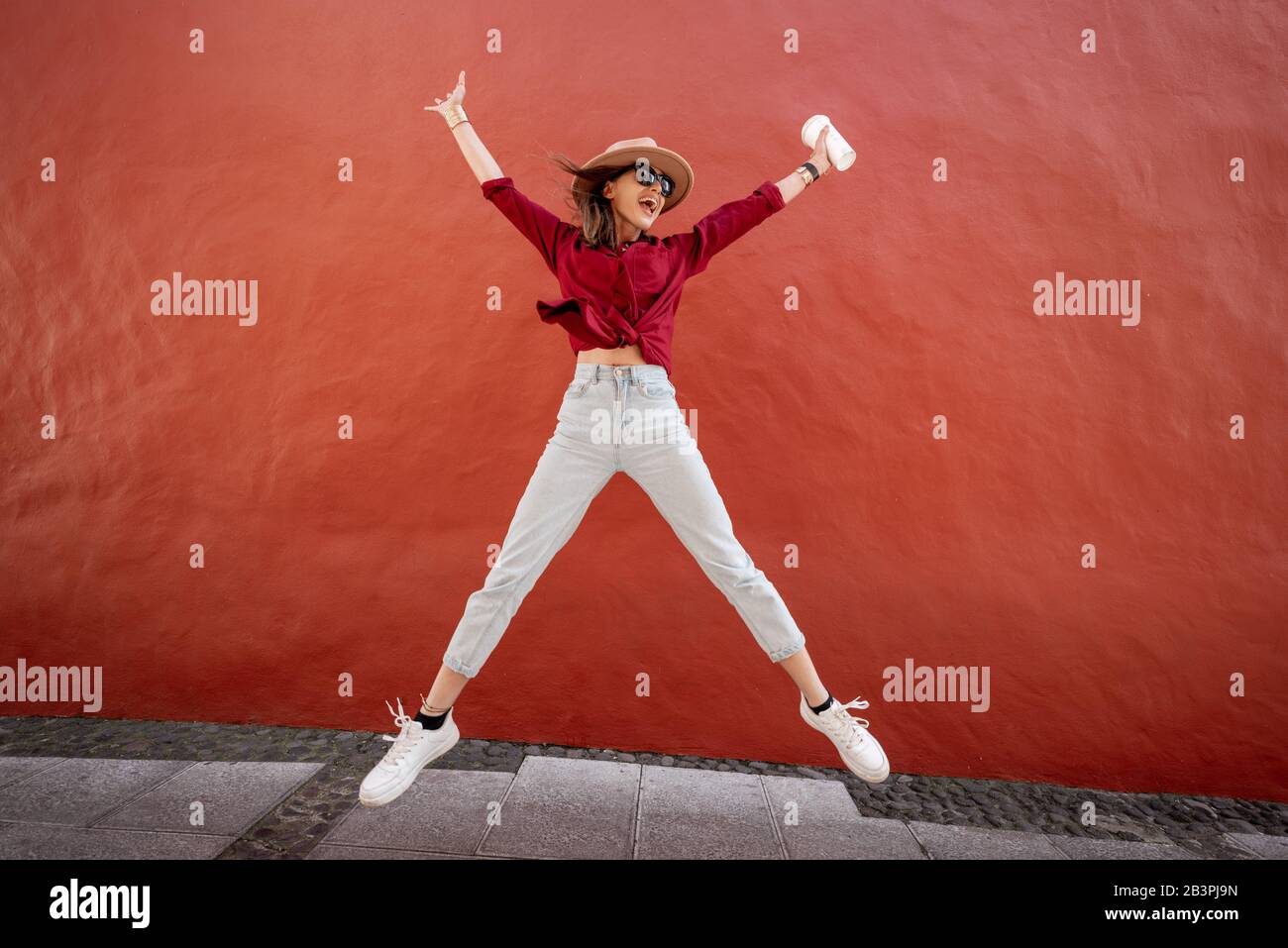 Portrait of an active stylish woman dressed in bright shirt with hat and coffee cup jumping on the red wall background outdoors. Carefree lifestyle, coffee and women's fashion concept Stock Photo