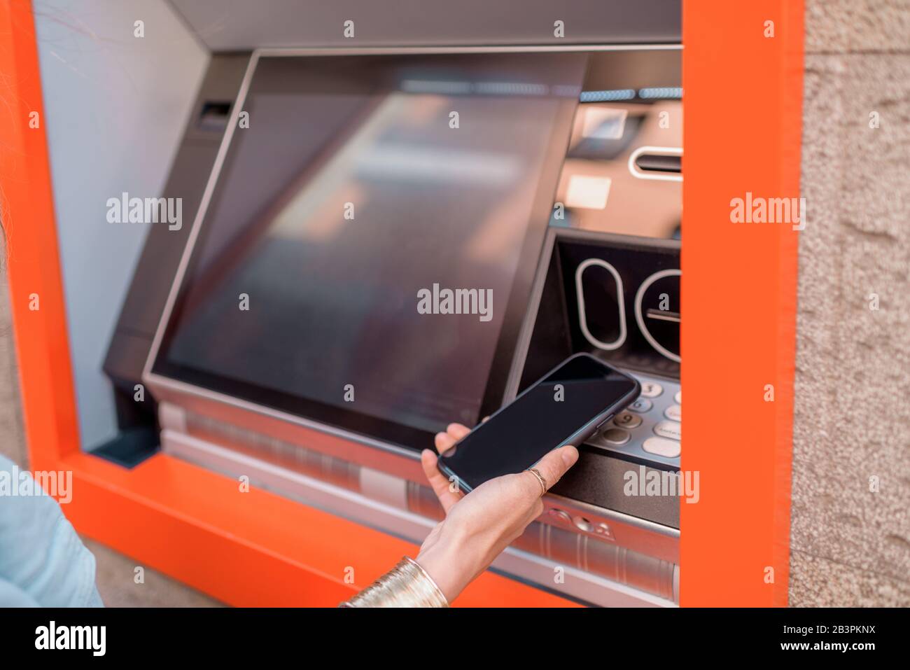 Young woman withdrawing cash from an ATM using a mobile phone on the street outdoors, close-up on phone Stock Photo