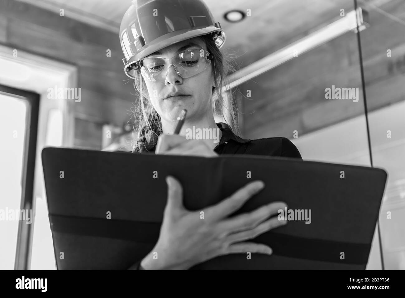 black and white low angle portrait of a woman holding a clipboard as she taking notes, writing reviews and reports. she wears construction hard hat. Stock Photo