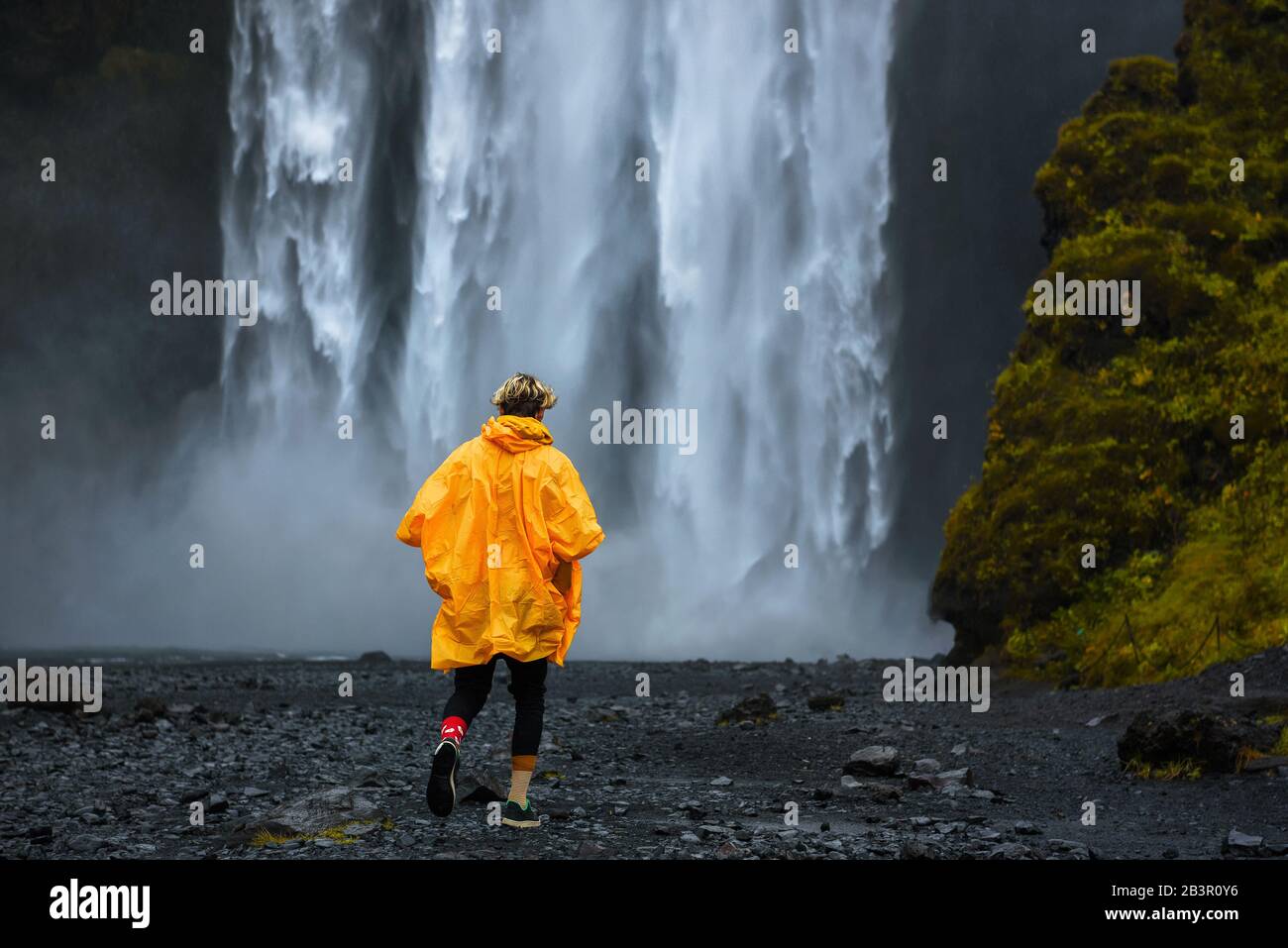 Tourist wearing a yellow raincoat walks to the Skogafoss waterfall in Iceland Stock Photo