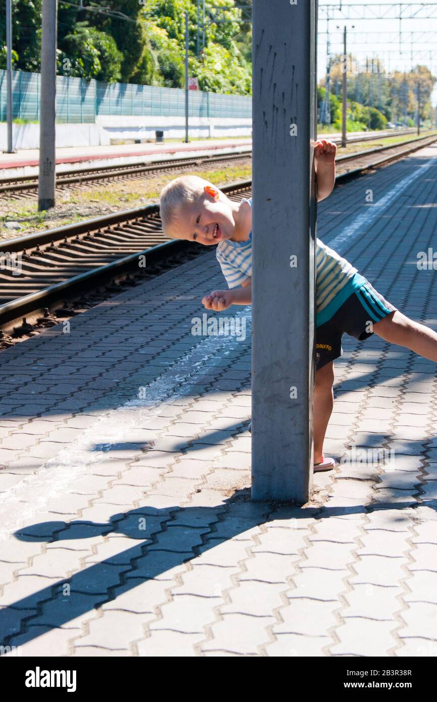 A little blond-haired boy of three years in a T-shirt and shorts on the platform is waiting for the train and playing catch-up - smiling, laughing, showing his tongue. Recreation, tropics, vacation, railway, station, summer Stock Photo