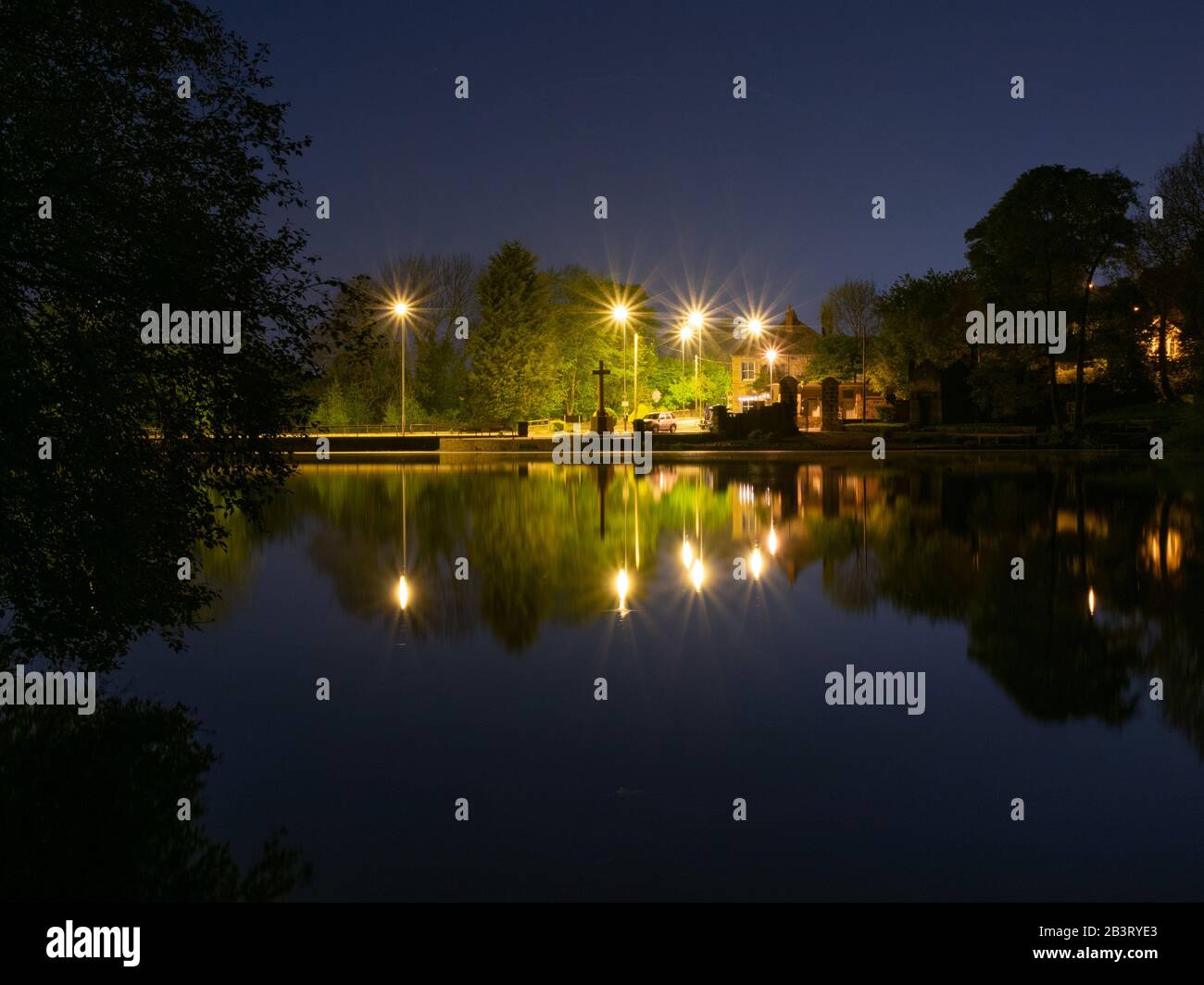 Street lights reflecting on a lake at midnight. With a war memorial in the centre, surrounded by foliage. With a clear dark blue sky. Stock Photo