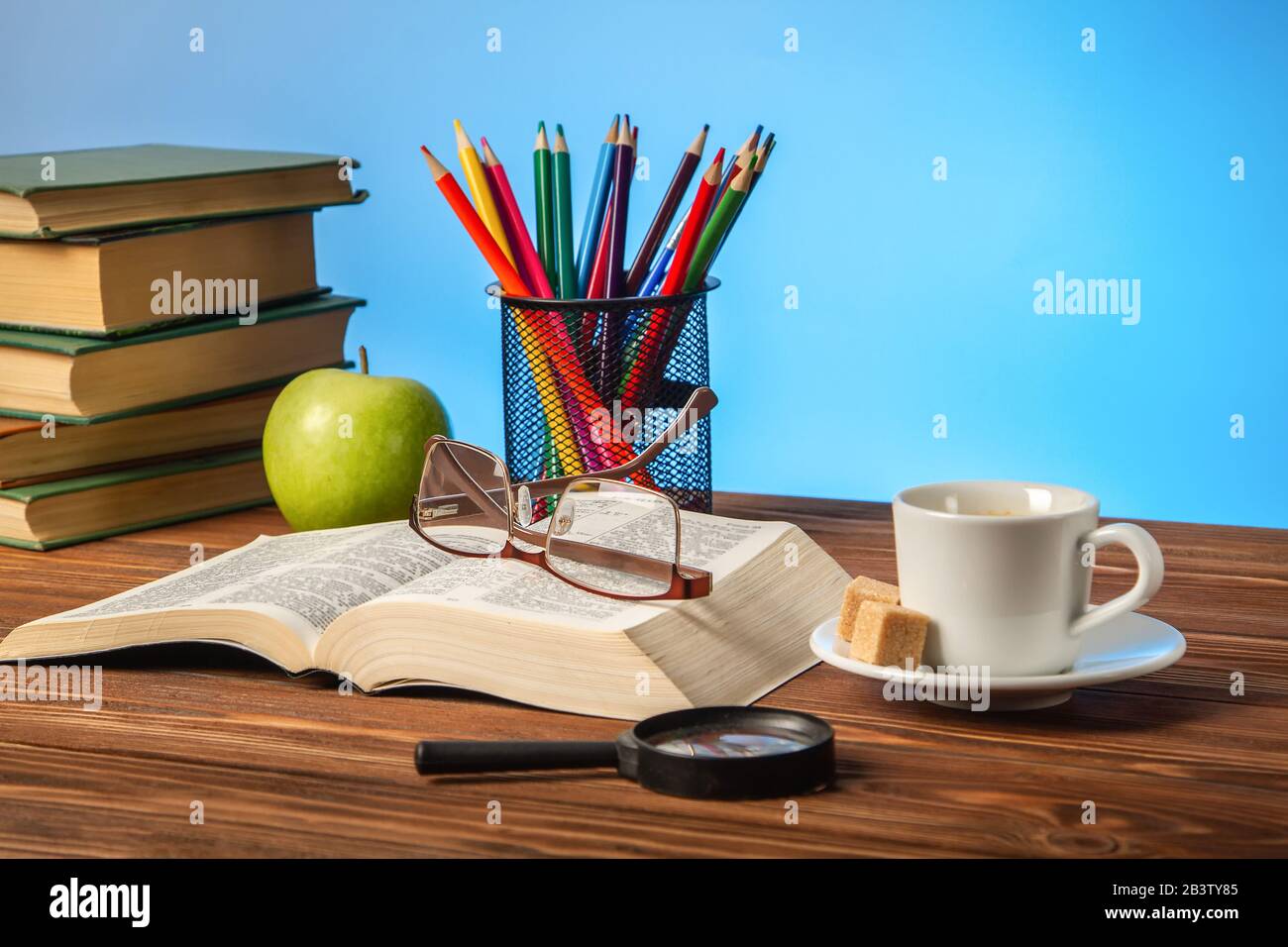 An open book on the table, glasses, a cup of cappuccino coffee with sugar cubes, color pencils on a blue background. Stock Photo
