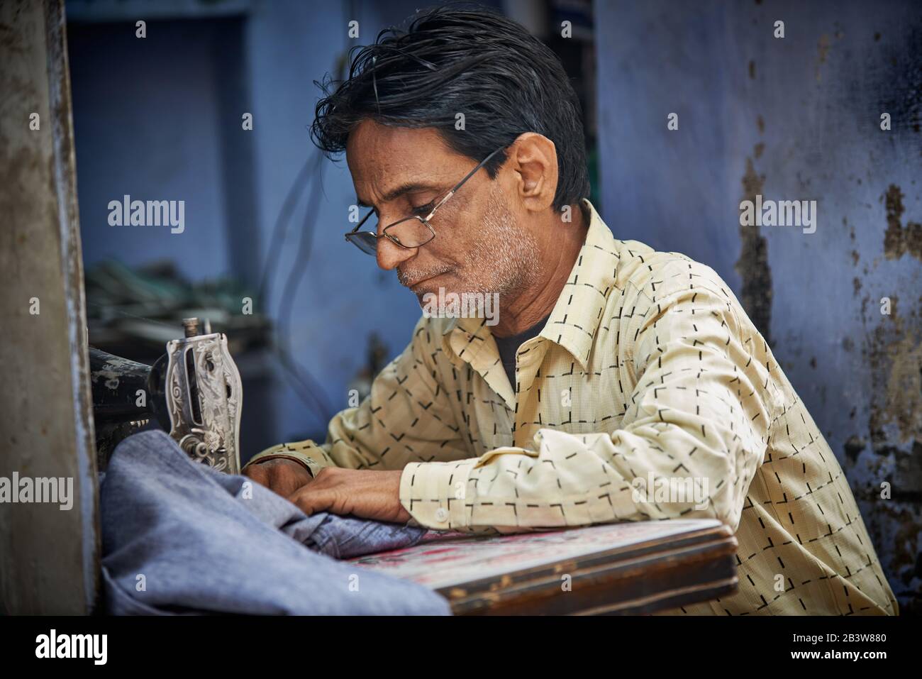 Portrait of a tailor on his sewing machine against a blue background, Bikaner, Rajasthan, India Stock Photo