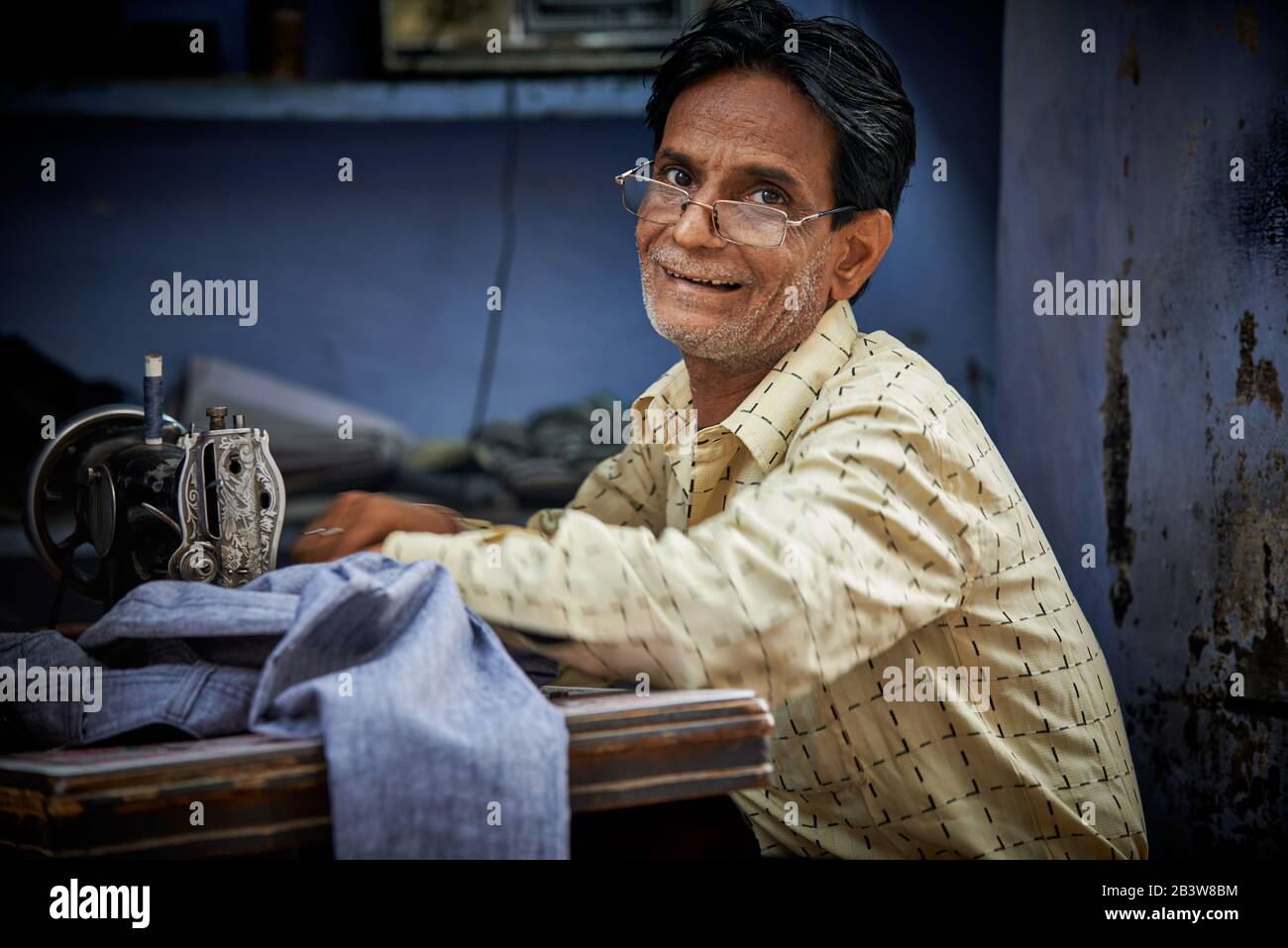Portrait of a tailor on his sewing machine against a blue background, Bikaner, Rajasthan, India Stock Photo