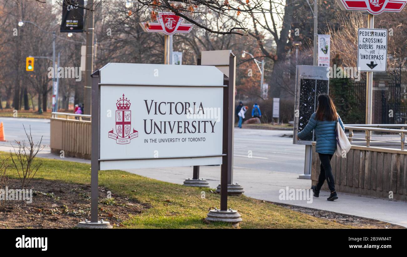 Victoria University (at the University of Toronto) logo on a sign out-front of campus in downtown Toronto as a women walks by the sidewalk. Stock Photo
