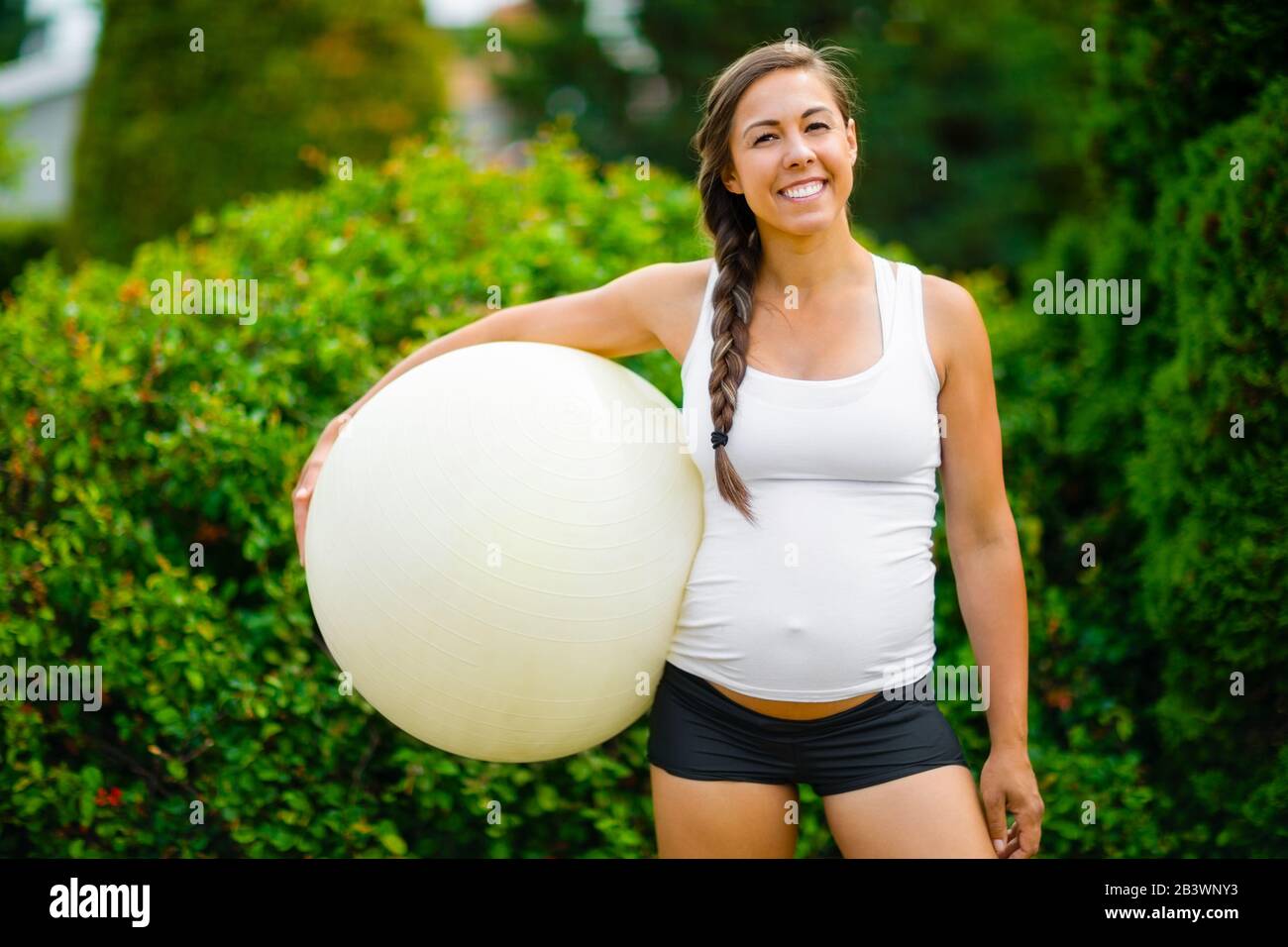 Smiling Young Expectant Mother Holding Exercise Ball In Park Stock Photo