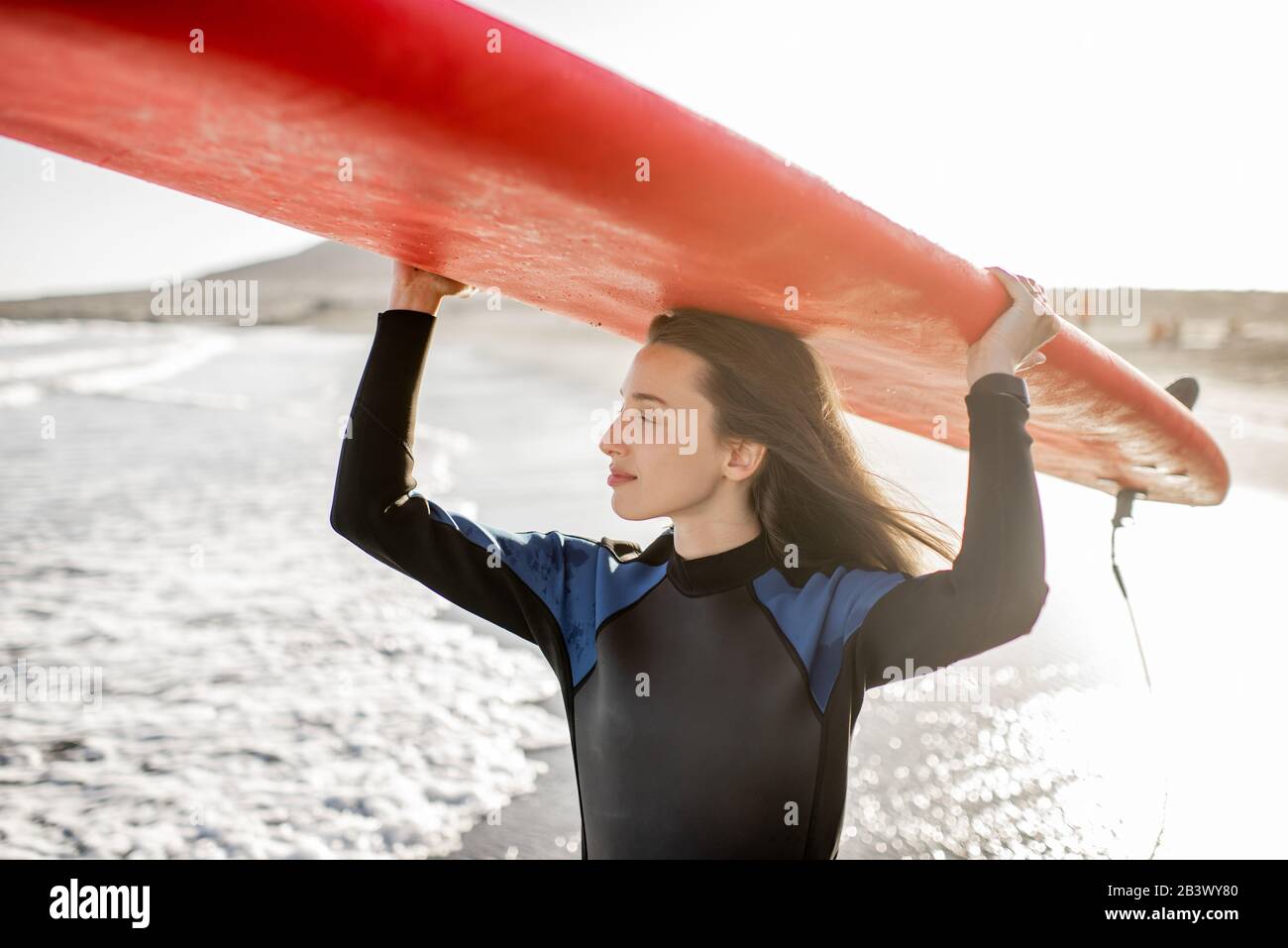 Portrait of a young woman in wetsuit carrying surboard above the head, preparing for surfing on the wild beach on a sunset. Active lifestyle concept Stock Photo