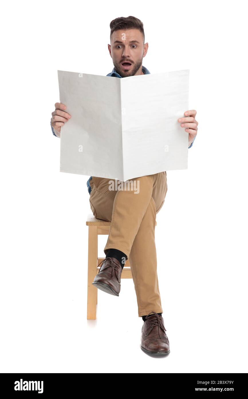 shocked young casual man reading newspaper and sitting isolated on white background, full body Stock Photo