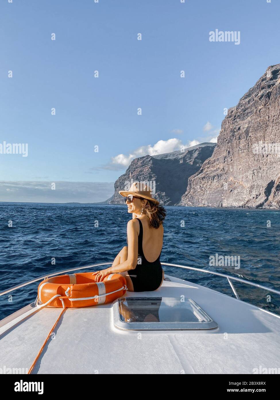 Woman enjoying ocean voyage sitting with lifebuoy on the yacht nose while sailing near the breathtaking rocky coast on a sunset. Image made on mobile phone Stock Photo