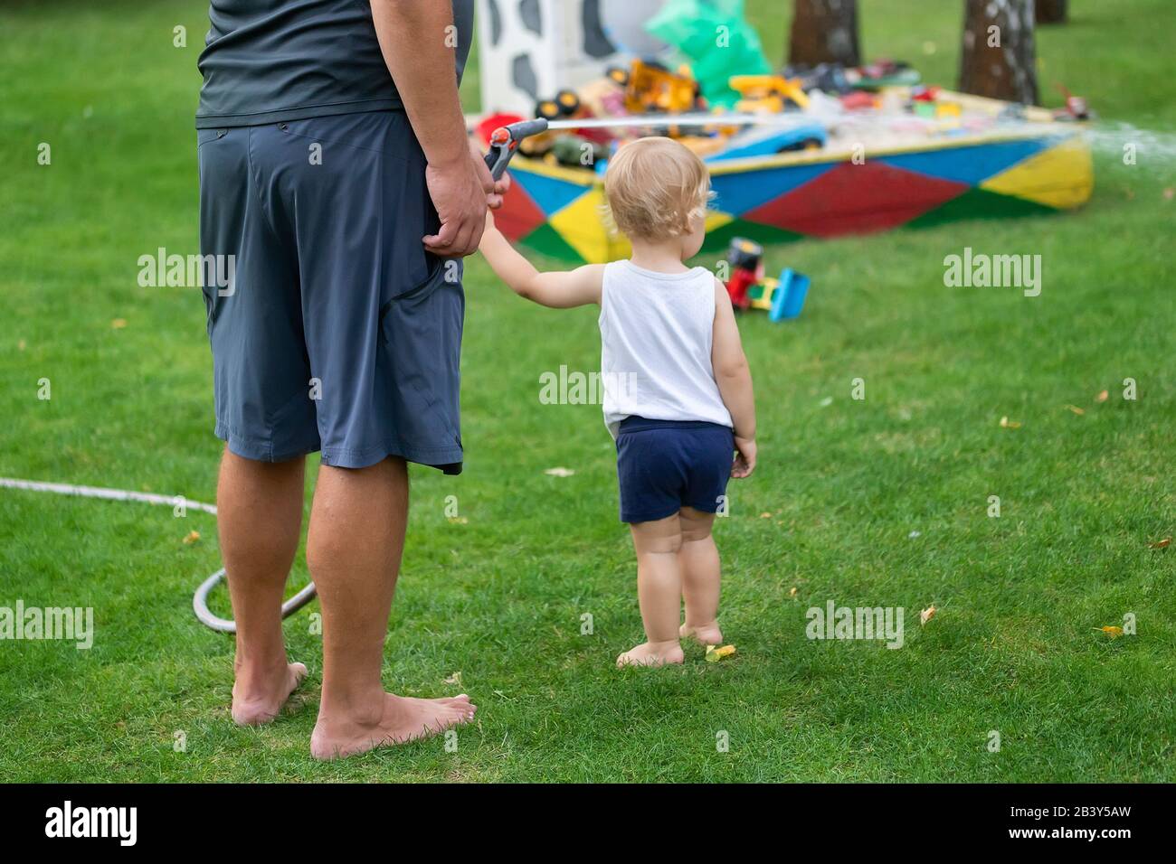 Father with son together watering garden and green grass lawn at home backyard outdoors on brigh summer warm day. Male person with little toddler boy Stock Photo